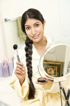 A young woman wears a bathrobe while holding a makeup brush and smiling at the camera. Her long dark hair is braided and hanging over her shoulder. Vertical shot.