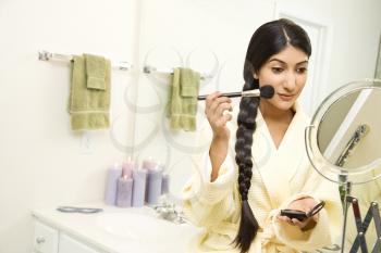 A young woman wearing a bathrobe is sitting on the sink in front of a mirror and applying makeup. Her long dark hair is braided and hanging over her shoulder. Horizontal shot.