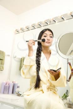 Young woman wearing a bathrobe is sitting on the sink in front of a mirror and applying makeup. Her long dark hair is braided and hanging over her shoulder. Vertical shot.