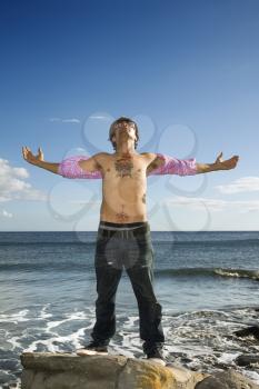 A tattooed young man standing on a rock in the ocean. He is looking up to the sky, and his arms are outstretched. Vertical shot.