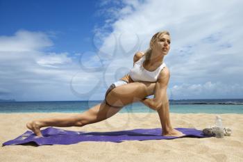 Attractive young woman stretches while doing yoga. She is on a sandy beach with the ocean in the background. Horizontal shot.