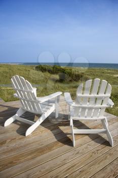 Two Adirondack style chairs sitting on a wooden deck, facing the shore. The grassy beach is in view beyond the deck. Vertical shot.