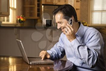 Man sitting at table smiling talking on cell phone and typing on laptop.