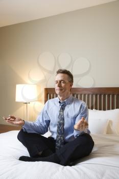 Caucasian businessman sits on a bed in the lotus position while meditating with closed eyes. Vertical shot.