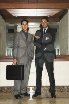 Young adult Asian and African-American businessmen standing in an office lobby looking at the camera. Vertical format.