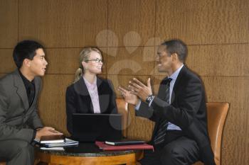 Three diverse businesspeople sit at a small table with a laptop and talk together. Horizontal format.