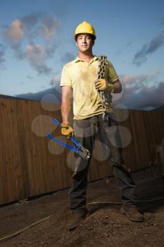 Attractive male construction worker in a hardhat stands at a construction site with bolt cutters and a chain. Vertical shot.