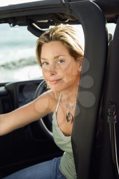 Portrait of a young Caucasian female sitting in the driver's seat of an SUV and smiling at the camera, with ocean waves in the background. Vertical format.