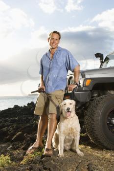 A smiling man leans against his SUV at the beach as he holds his dog's leash and looks at the camera. Vertical format.