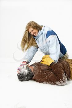 Young woman playfully sits atop a young man and hits his head with a snowball. Vertical shot.