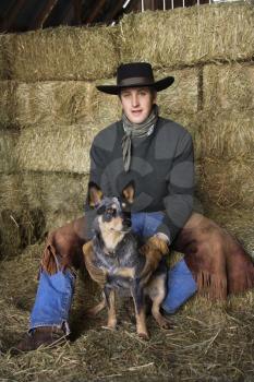Attractive young man wearing a black cowboy hat, sitting atop a hay bale. He is smiling and holding an Australian Shepherd. Vertical shot.