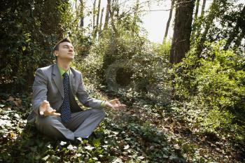 Young businessman sits in a lotus position meditating in the woods as sunlight filters in through the trees. Horizontal shot.