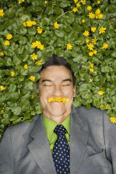 Young smiling businessman relaxing in a flower patch wearing flowers over his mouth like a mustache.
