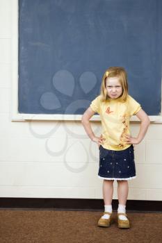 Girl standing in school classroom with hands on hips and pouty facial expression. Vertically framed shot.