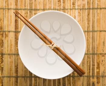 Overhead view of chopsticks lying across an empty bowl on a bamboo mat. Horizontal format.