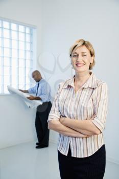 Smiling professional businesswoman standing as man reads architectural plans in background.