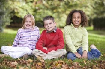 Royalty Free Photo of Three Children Sitting on the Grass With Autumn Leaves in Front of Them