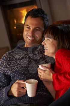 Middle Aged Couple Sitting On Sofa By Cosy Log Fire With Hot Drink