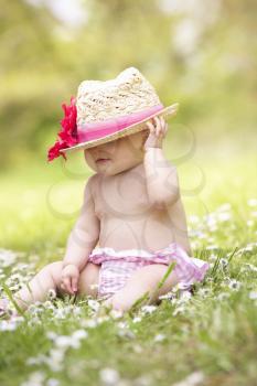 Baby Girl In Summer Dress Sitting In Field Wearing Straw Hat