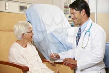 Doctor Taking Notes From Senior Female Patient Seated In Chair By Hospital Bed