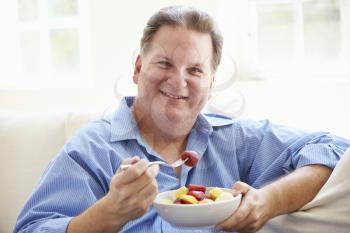 Overweight Man Sitting On Sofa Eating Bowl Of Fresh Fruit