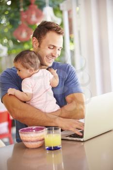 Father Holding Baby And Making Snack Whilst Using Laptop