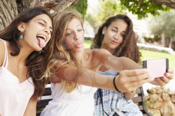 Three Teenage Girls Sitting On Bench Taking Selfie In Park