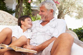 Grandfather And Grandson Sitting In Garden Reading Book