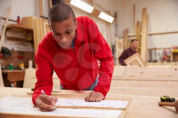 Apprentice Working With Plans In Carpentry Workshop