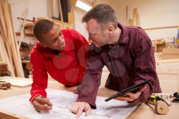 Carpenter With Apprentice Looking At Plans In Workshop