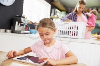 Girl Using Digital Tablet As Mother Sorts Laundry