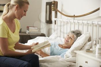Adult Daughter Reading To Senior Female Parent In Bed At Home