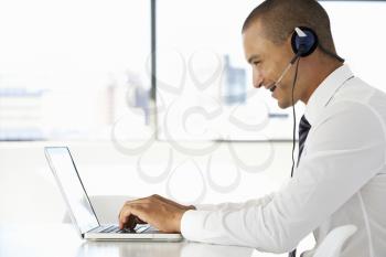Businessman Sitting At Desk In Office Using Laptop Wearing Headset