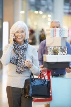 Senior Woman Shopping In Mall As Husband Carries Boxes