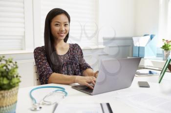 Female Doctor Sitting At Desk Working At Laptop In Office