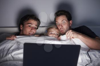 Young girl and two dads watch scary film in bed on laptop