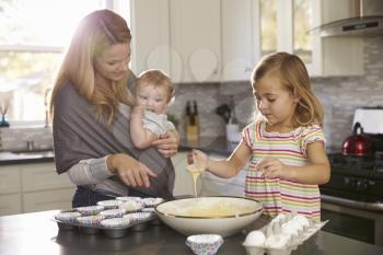 Young girl preparing cake mix in kitchen, mum showing baby