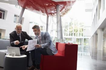 Two Businessmen Discuss Document In Lobby Of Modern Office