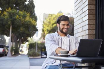 Bearded young man using a laptop at a table outside a cafe