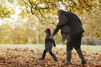 Grandfather And Granddaughter Kicking Leaves On Autumn Walk