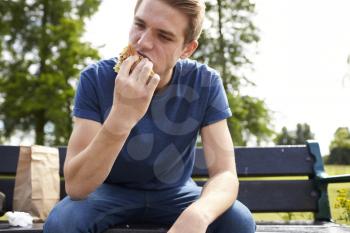 Young Man Sitting On Park Bench Eating Burger