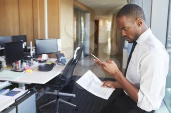 Businessman At Desk Checking Messages On Mobile Phone