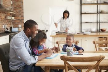Father And Children Drawing At Table As Mother Prepares Meal