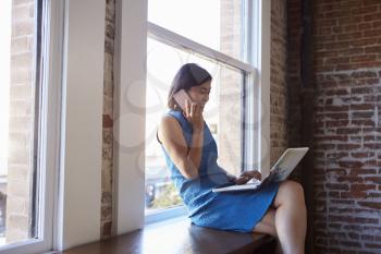 Businesswoman By Window Using Mobile Phone And Laptop