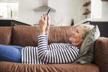 Senior white woman lying on couch at home using smartphone
