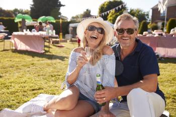 Portrait Of Mature Couple Sitting On Rug At Summer Garden Fete With Drinks