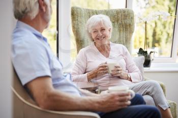 Senior Couple Sitting In Chairs And Chatting In Retirement Home