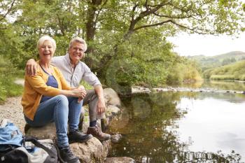 Portrait Of Senior Couple On Hike Sitting By River In UK Lake District