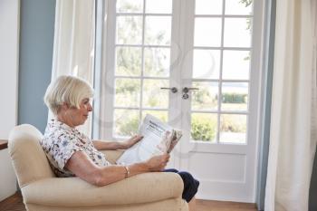 Senior woman sitting in armchair reading a newspaper at home