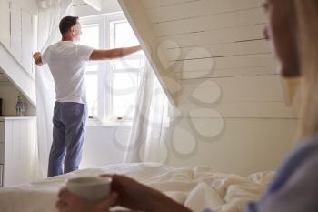 Man Opens Curtains And Looks Out Of Window As Woman Lies In Bed